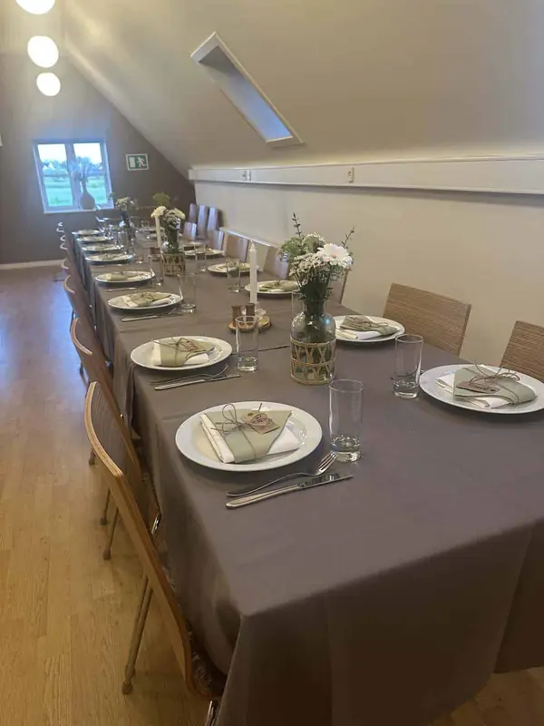 Langt bord under skråt loft med grå dug, grønne servietter og hvide blomster